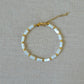 A delicate gold chain bracelet with alternating white rectangular beads and small gold spacer rings, displayed on a textured beige linen background.