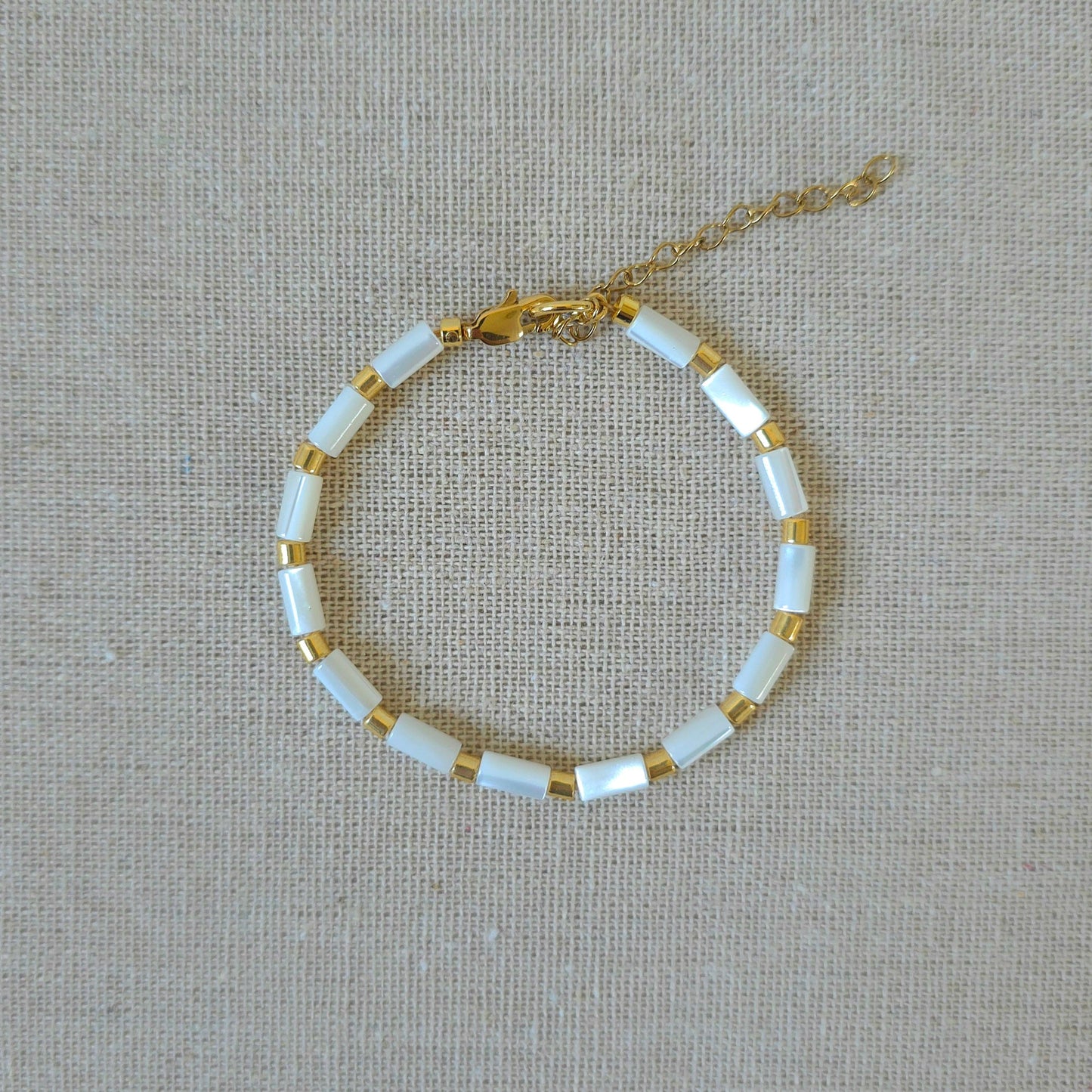 A delicate gold chain bracelet with alternating white rectangular beads and small gold spacer rings, displayed on a textured beige linen background.